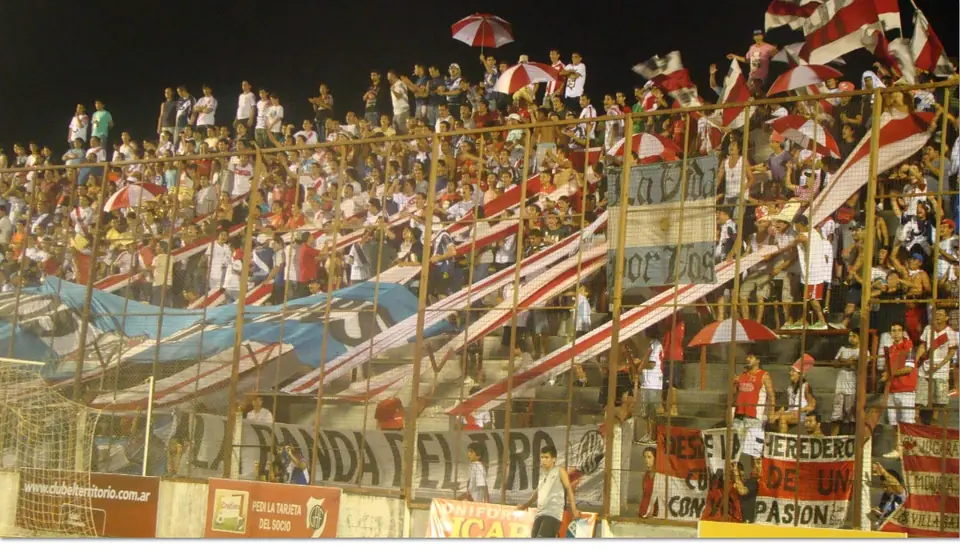 La tribuna colmada de público, con banderas pintadas a mano colgadas del alambrado.