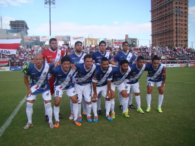 Un plantel formado en el campo antes de un partido, con la tribuna y la ciudad detrás.